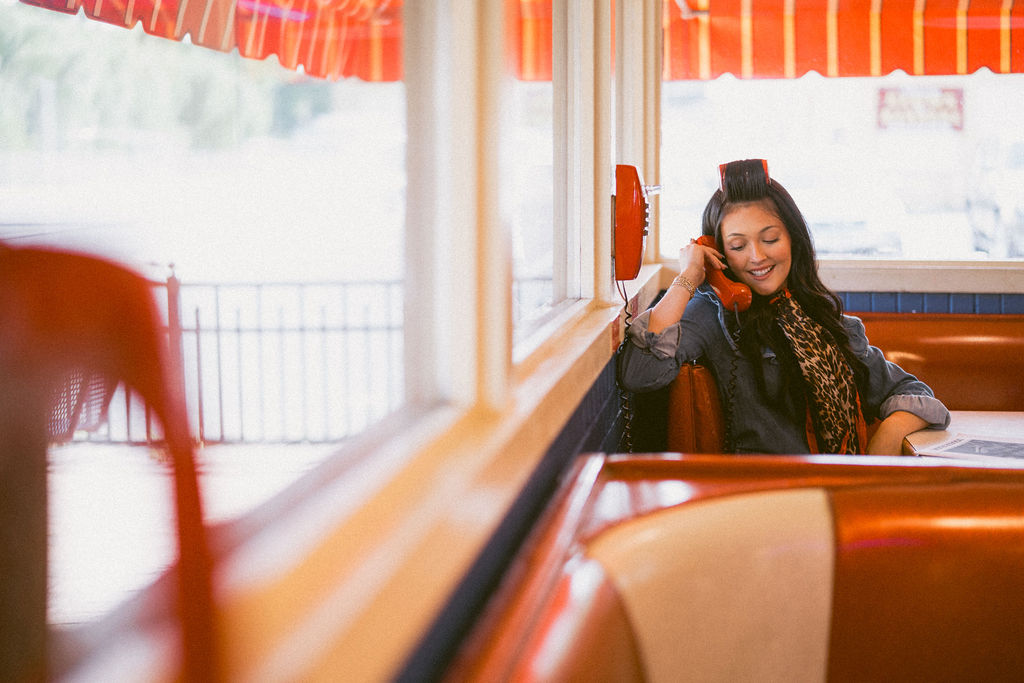 Hairstylist smiling with a vintage red telephone in a colorful diner booth.