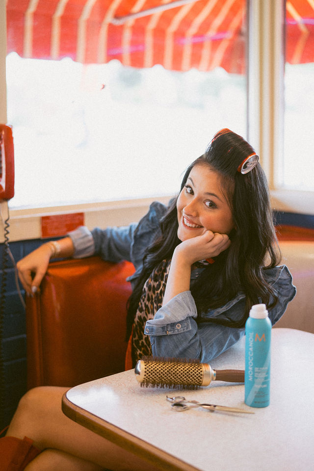 Hairstylist leaning on the table in a diner booth with a hair roller in place.