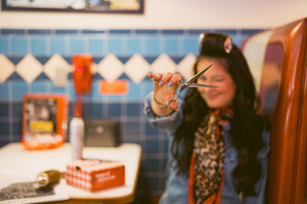 Hairstylist holding a pair of scissors toward the camera in a diner booth.