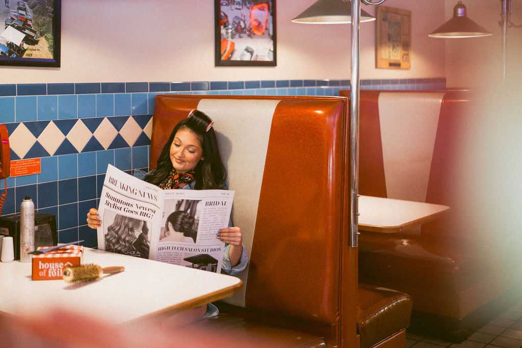 Hairstylist reading a branded newspaper in a warm orange diner booth.