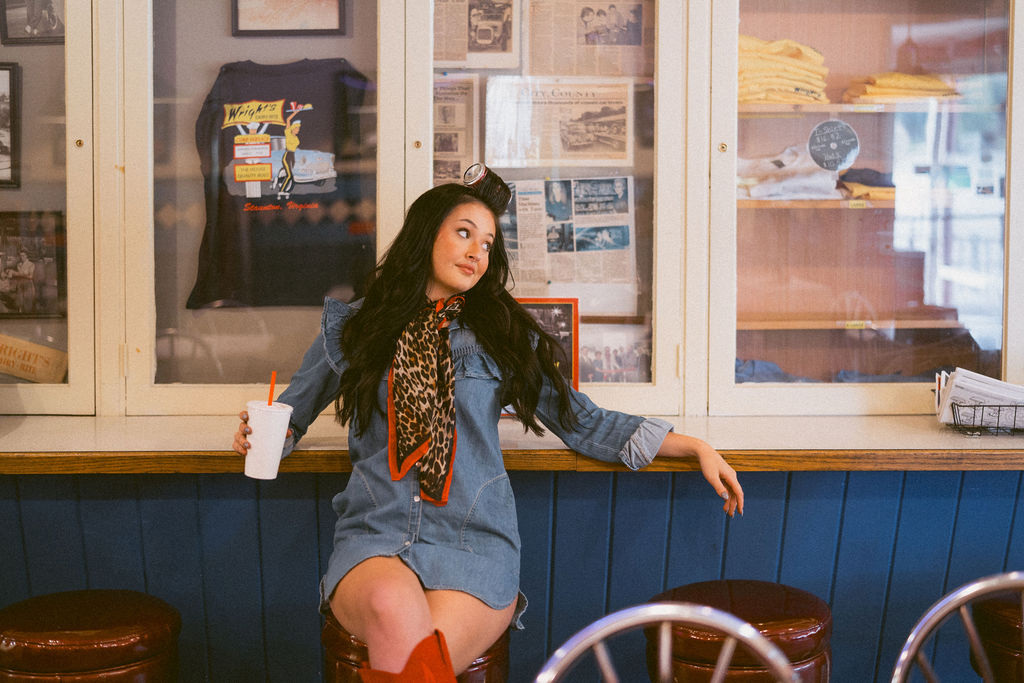Hairstylist seated at a diner counter with a drink and framed photographs behind her.
