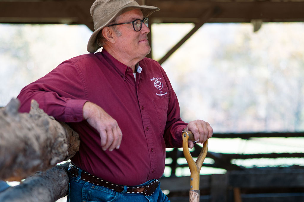 Farmer in a burgundy shirt and wide-brimmed hat leaning on a wooden barn rail.
