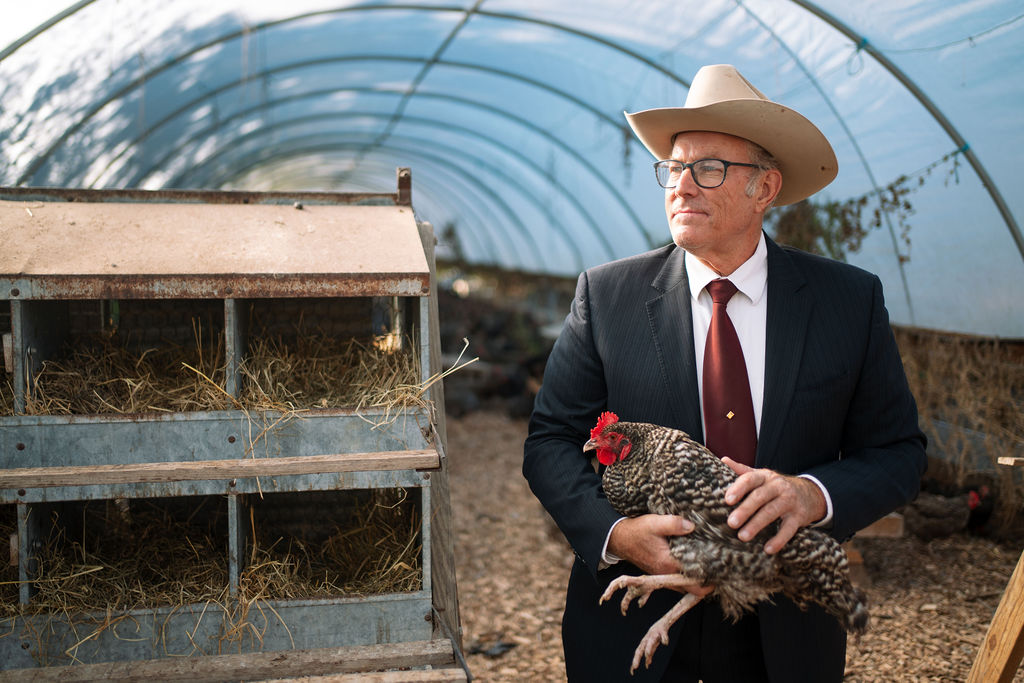 Joel Salatin in a dark suit, tie, and cowboy hat holding a chicken inside a Polyface Farm hoop-house.