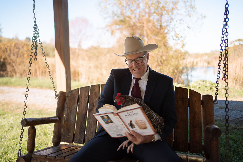 Joel Salatin in a suit and cowboy hat reading on a porch swing with a chicken in his lap.